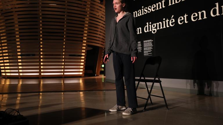 A young woman talks in front of a black wall that says, in large white letters, “Tous les êtres humains naissent libres et égaux en dignité et en droits.” (All human beings are born free and equal in dignity and rights.) There is a circular theatre in the background that resembles a large woven basket. Partially obscured.