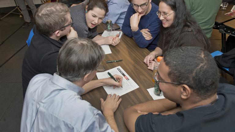 A group of excited people gathered around a table where one man is writing on a piece of paper. Partially obscured.