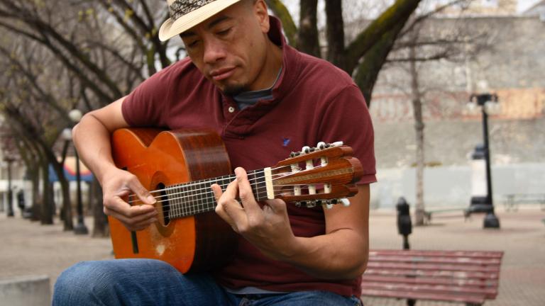 A man wearing a fedora plays a guitar while sitting outside on a bench. There are trees in the background. Partially obscured.