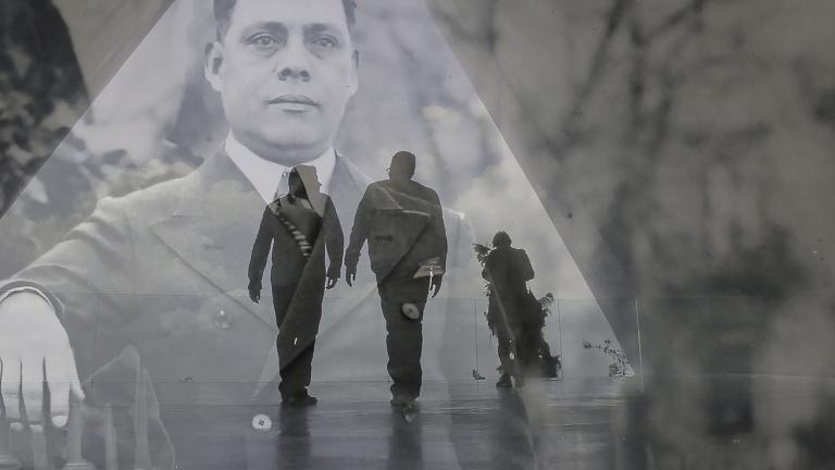 A translucent historical image of a man in a suit layered over a contemporary photo showing the dark outline of two men walking in a hallway. Partially obscured.