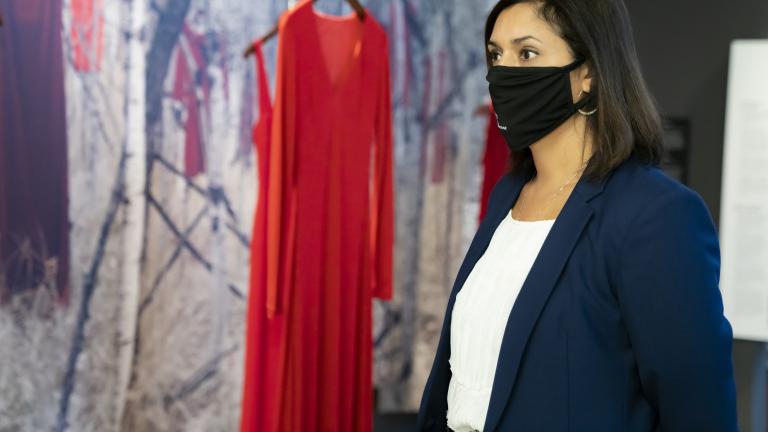 A woman wearing a black face mask stands in front of a museum exhibit consisting of red dresses hanging in front of a woodland background. Partially obscured.