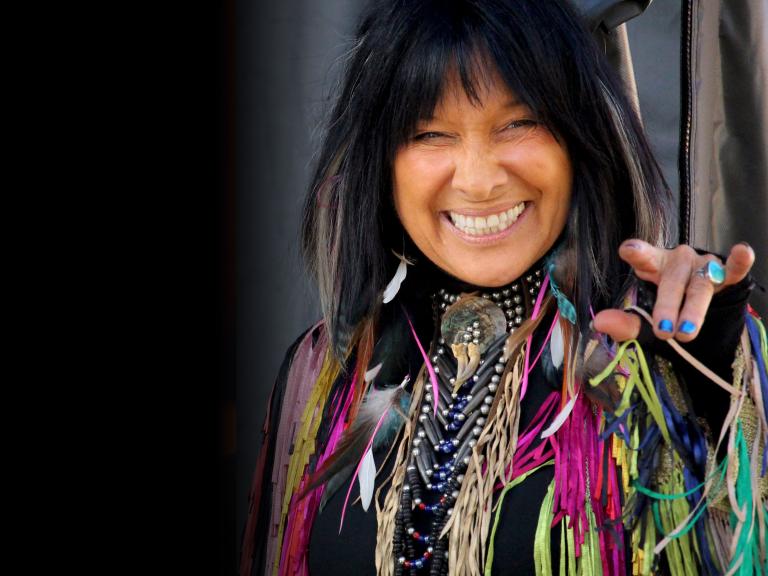 Indigenous woman with dark hair smiles and is highly decorated with beads and many colourful ribbons. Partially obscured.