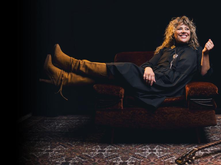 A very happy woman with curly blond hair and a guitar at her feet is sitting with her feet up on a couch. Partially obscured.