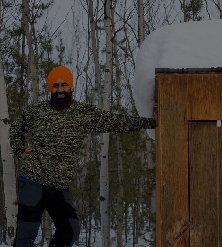 A Sikh-Canadian man, smiling, wearing a green sweater, blue and brown pants, boots, and an orange turban, leaning against a cabin with a crescent moon. Partially obscured.