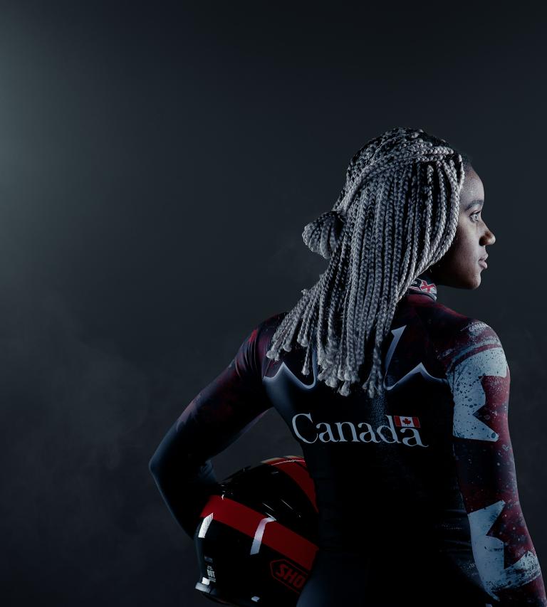 A black woman stands in a dark place between two large projectors. She's holding a helmet and wears a Canadian bobsleigh suit. Partially obscured.