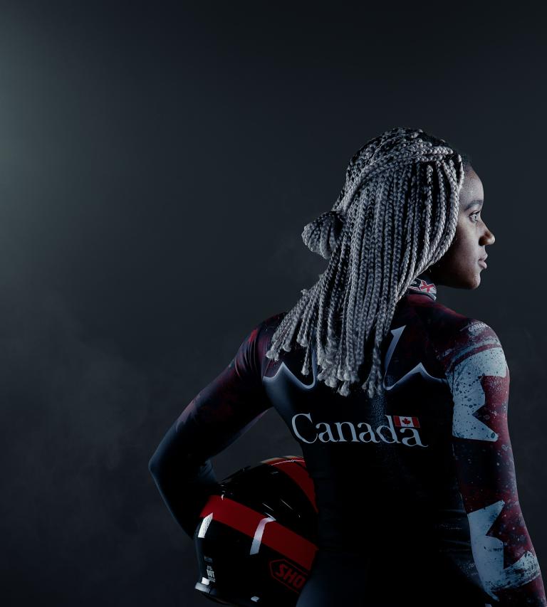 A black woman stands in a dark place between two large projectors. She's holding a helmet and wears a Canadian bobsleigh suit.