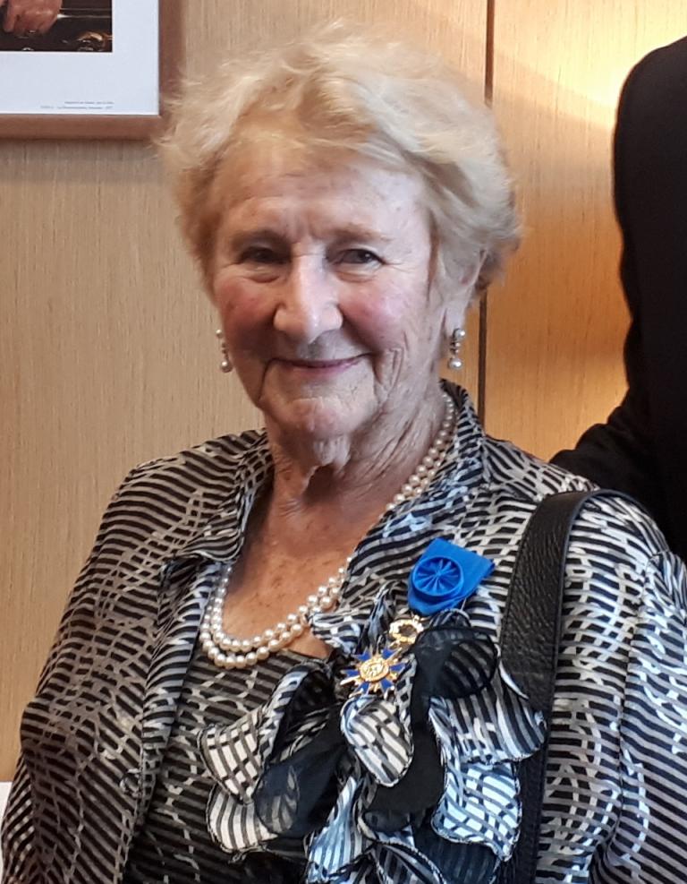 An elderly woman with short gray hair smiles gently at the camera. She is wearing pearl earrings, a pearl necklace, and a patterned black-and-white blouse. She stands in warm light in front of a wood-panelled wall. Partially obscured.