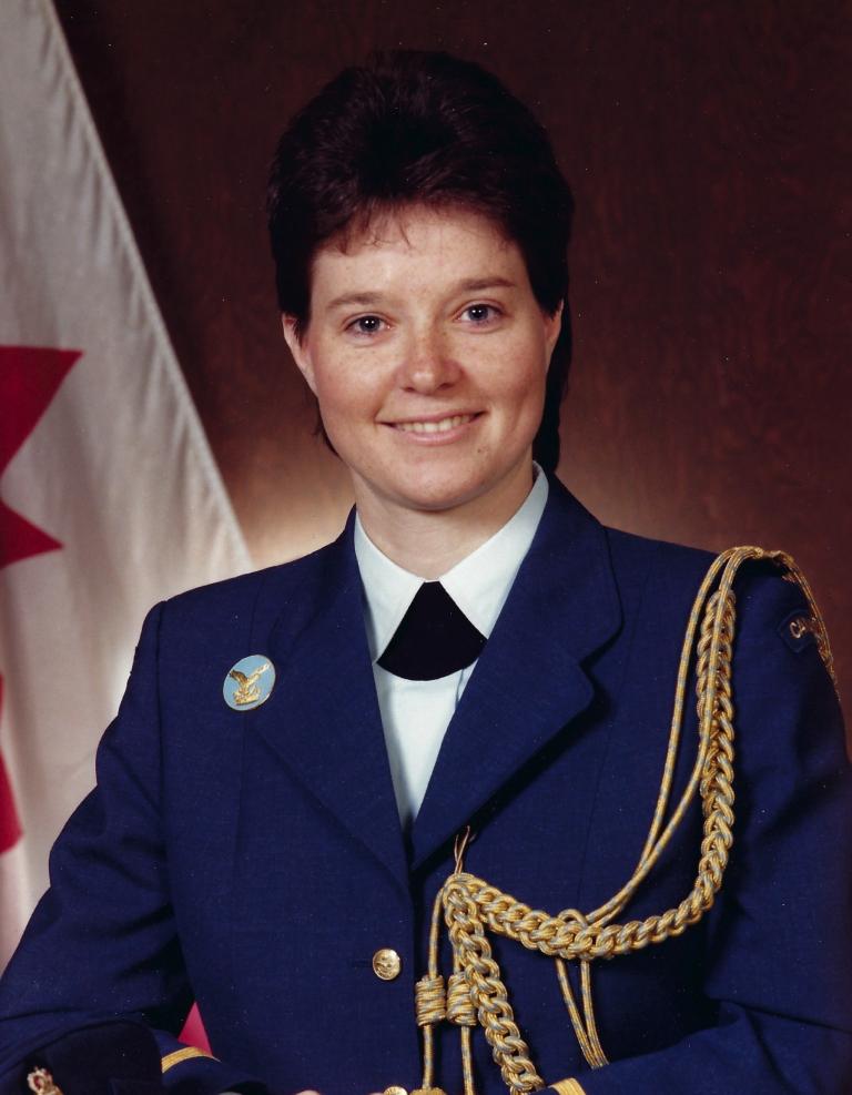  A photograph shows a smiling young woman with short brown hair wearing a blue jacket with gold trim and a short black scarf over a white shirt. Partially obscured.