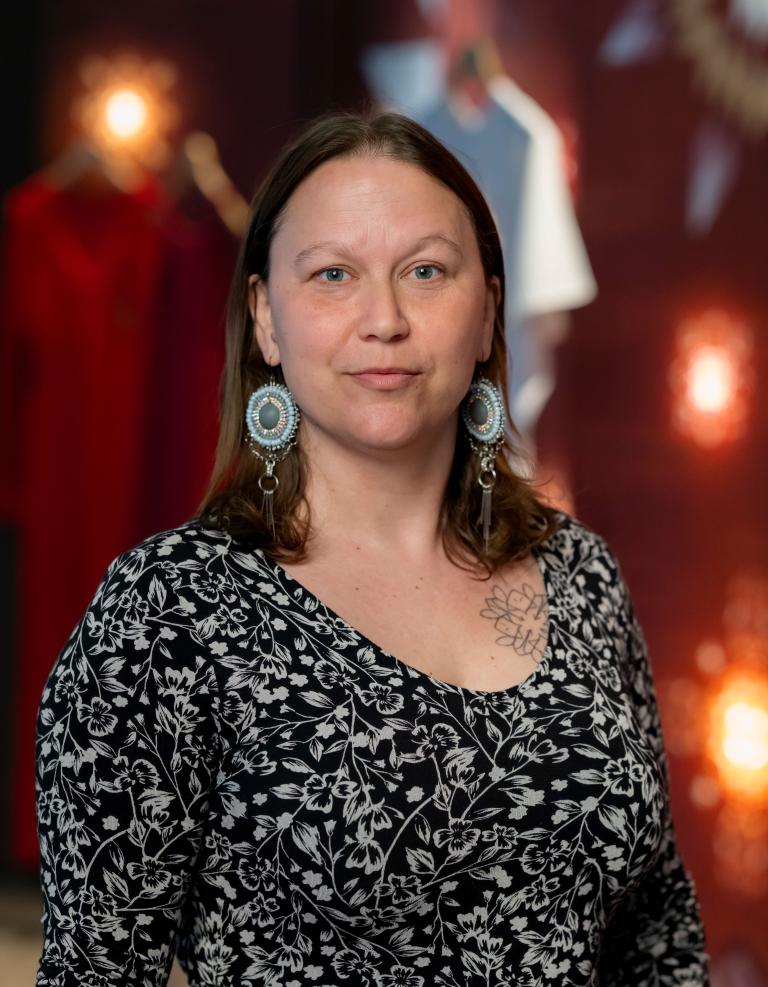 A woman stands proudly in front of red dresses in the background. She wearing a black and white floral dress and ornate light blue circular earrings. Partially obscured.