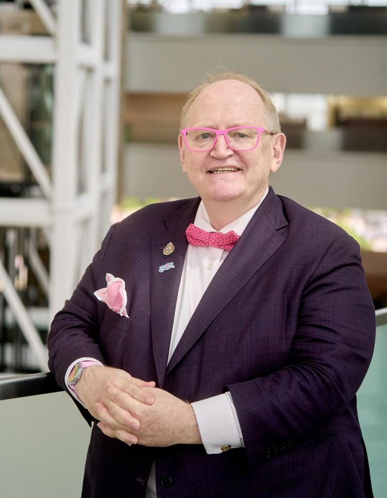 A smiling older gentleman wearing pink-framed glasses, a purplish suit and a pink polka-dot bowtie. Partially obscured.