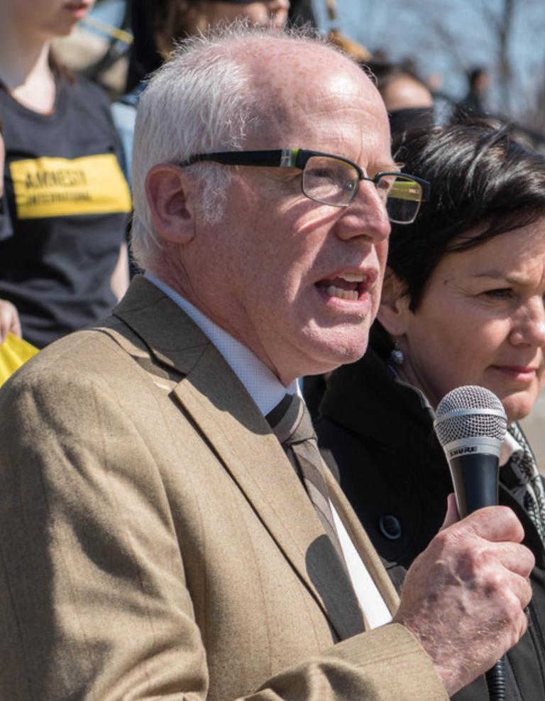 A middle-aged man with short grey hair stands with a mic speaking to a crowd. Partially obscured.