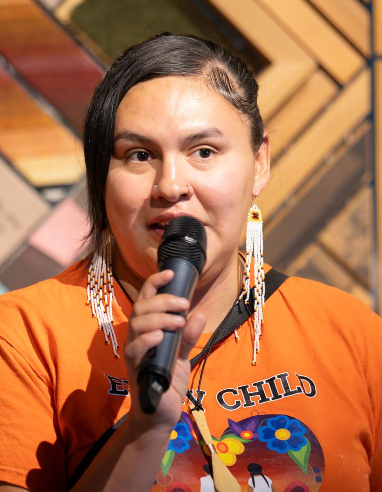 A dark-haired Indigenous person wears long beaded earrings and an orange shirt that reads "Every Child Matters." They are speaking into a microphone and standing in front of a wooden artpiece that has many different coloured segments. Partially obscured.