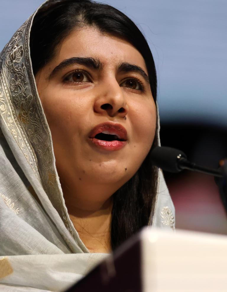  Malala Yousafzai, a young woman wearing a light-coloured hijab, is speaking in a microphone. Partially obscured.