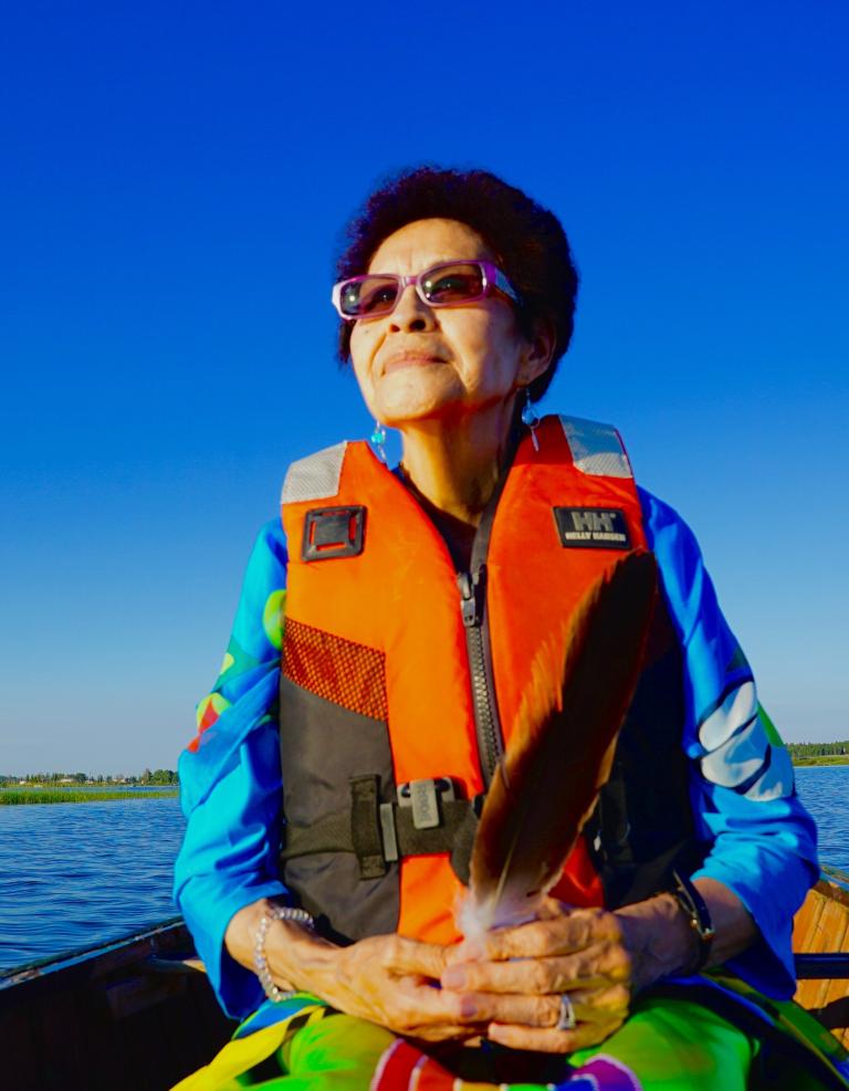 An indigenous woman is in a canoe on a lake with an orange life jacket. She seems at serene. Partially obscured.