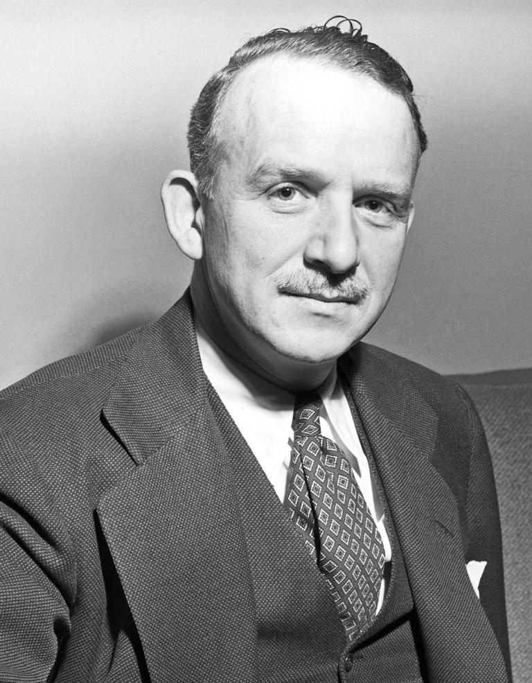 A black-and-white photograph of an older man sitting on a sofa. He is dressed in a suit with a patterned tie and vest, resting one arm on the armrest and looking calmly toward the camera with a slight smile. Partially obscured.