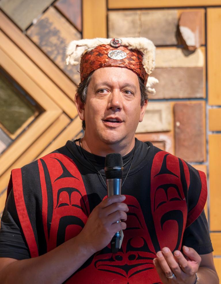 A man stands with a mic speaking to a crowd wearing traditional indigenous clothing infront of a wood panel art installation. Partially obscured.