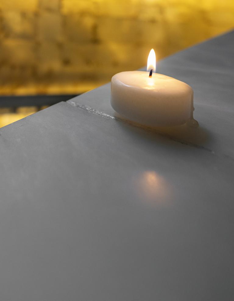 A small lit candle sits on a smooth stone surface in the foreground, its flame glowing softly. In the background, a long alabaster-lined corridor with handrails is illuminated by warm, ambient lighting, creating a quiet, reflective atmosphere. Partially obscured.