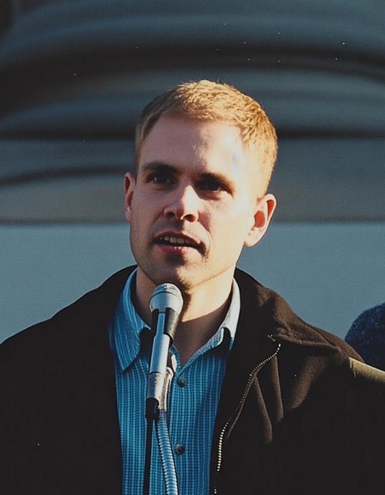 A young man with short blonde hair stands on the steps of a staircase of a courthouse speaking into a mic while someone takes a photo of him in the background. Partially obscured.