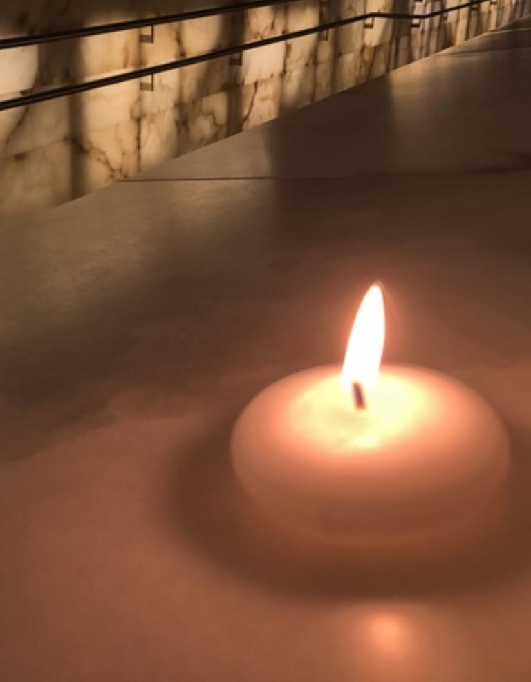 A small lit candle sits on a smooth stone surface in the foreground, its flame glowing softly. In the background, a long alabaster-lined corridor with handrails is illuminated by warm, ambient lighting, creating a quiet, reflective atmosphere. Partially obscured.