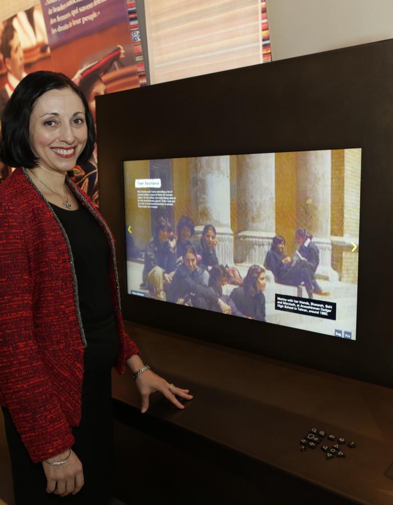 Marina Nemat smiles and stands beside a large digital display screen. She is wearing a red jacket and is looking directly at the photographer. The screen features a picture of people sitting on steps in front of large stone columns. 