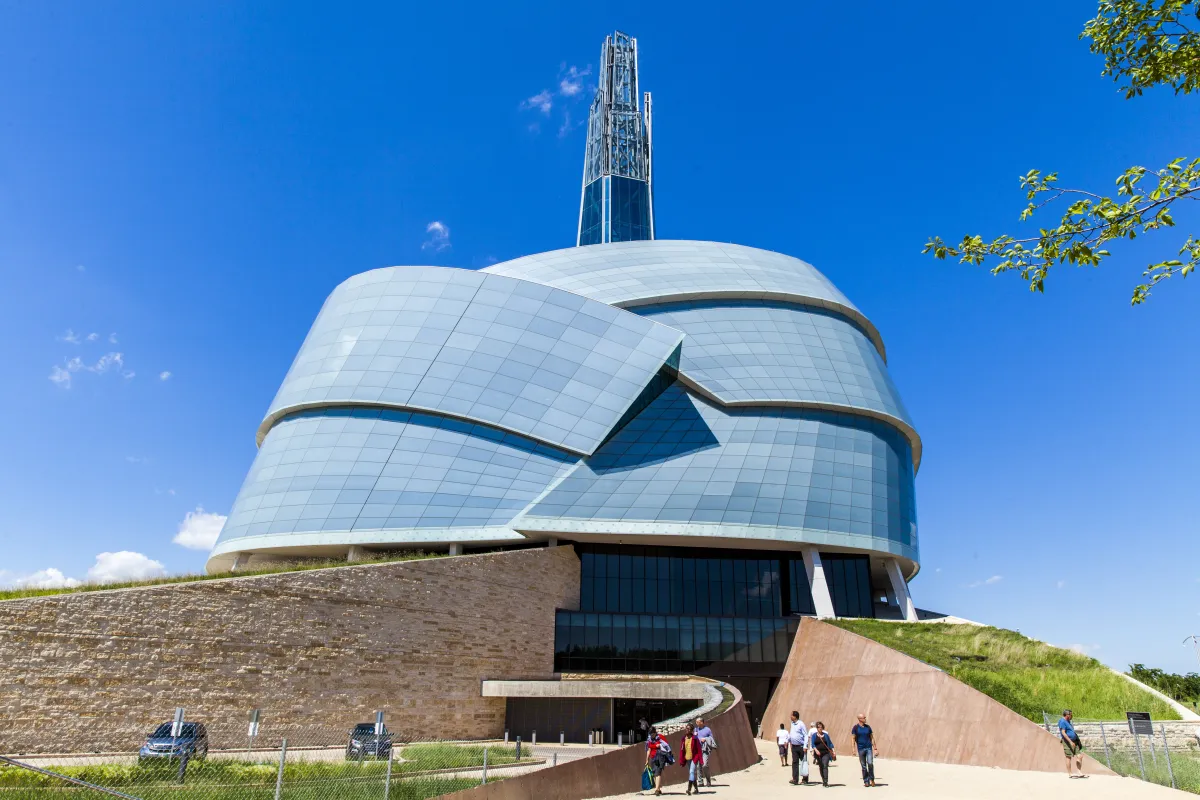 The Museum's entrance presents several ramp structures, some with grassy tops that extend all the way to the roof, which has a dome-shape formed by panes of glass resembling the folded wings of a dove. A tower rises in the center like a mountain peak. It is a sunny day with a bright blue sky and a small group of visitors are exiting the Museum.