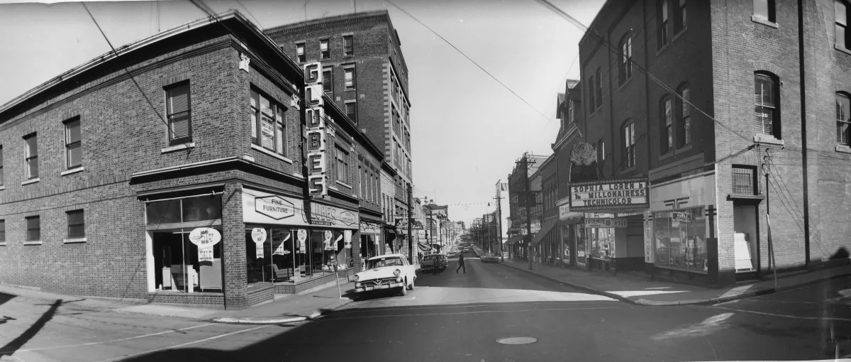 A black and white image of a town street. The street leads away from the viewer towards a vanishing point on the horizon, and is lined with brick buildings ranging from two to seven stories storeys tall. Many of the buildings have storefronts on the first floor, and the first building on the right has a movie marquee advertising a film called “The Millionairess” starring Sophia Loren.