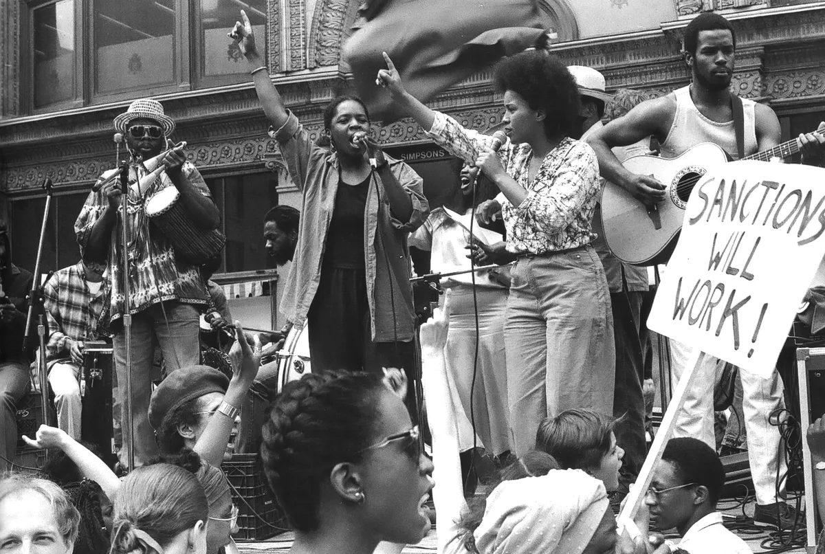 Men and women musicians on a stage. The two singers in the centre have their right arms raised. In the crowd watching them are some protest signs.