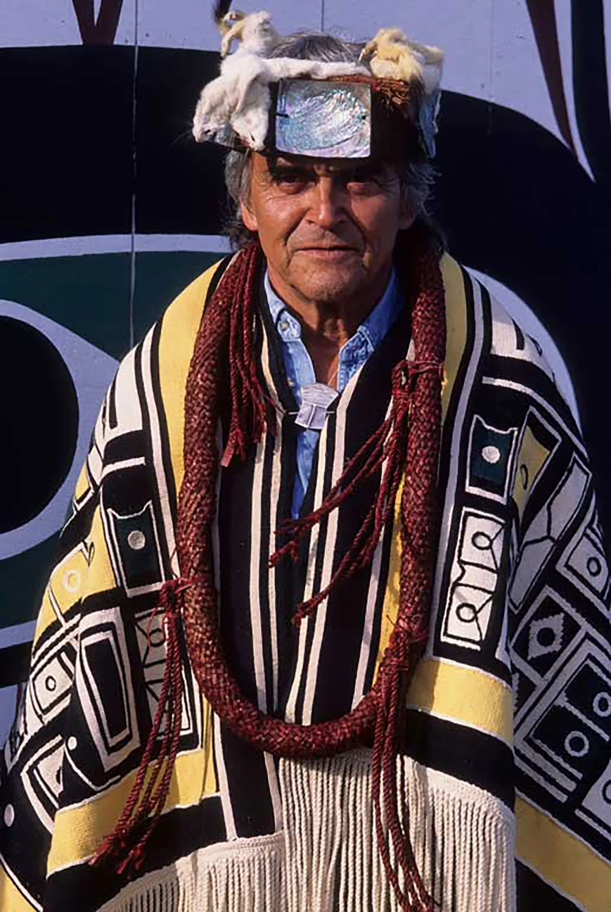 Bill Cranmer, an older man with gray hair, stands facing the camera. He is wearing a fringed blanket over his shoulders that decorated with geometric designs. He is also wearing fur hat with a shiny square metal centrepiece and a large necklace made out of a thick cord. 