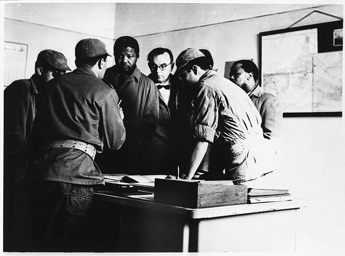 A black-and-white image of Nelson Mandela standing in front of a desk. He is surrounded by a group of five men, some of whom are wearing military clothing. One man, with his back to the viewer, appears to be speaking to Mandela, while the other men are looking at something on the desk.