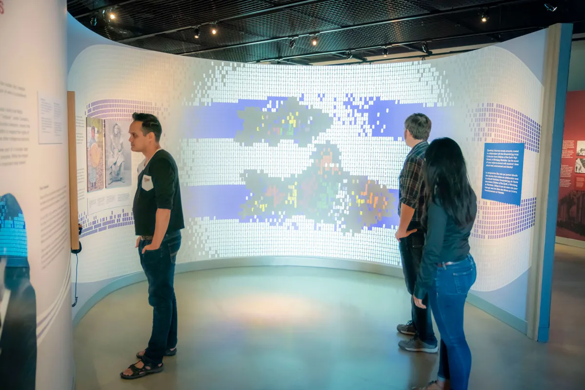 Three people stand in front of an image being projected onto a curved wall that is about nine feet tall. Two are looking at the image, while third is to the viewer’s left, at something else on the wall. The image on the wall is made up of many small squares and has the appearance of Indigenous bead work or a mosaic. The image is mainly made up of white squares, but it also features two thick blue horizontal lines that run parallel to each other. Each line is broken up near its centre by a representation of 