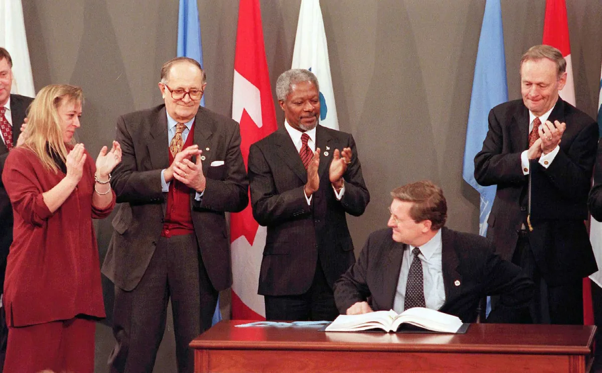 One man is sitting at a table in front of an open book. One woman and three men are standing behind him and applauding.