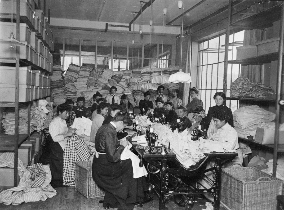 A large group of women sitting and sewing. Others are standing behind them in a room crowded with textiles. 