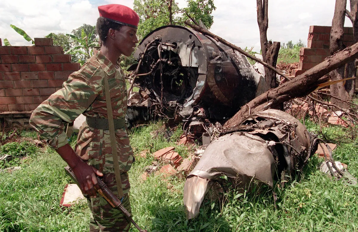 A soldier carrying a rifle and wearing a red beret and camouflage uniform stands in front of the wreckage of a plane among trees.