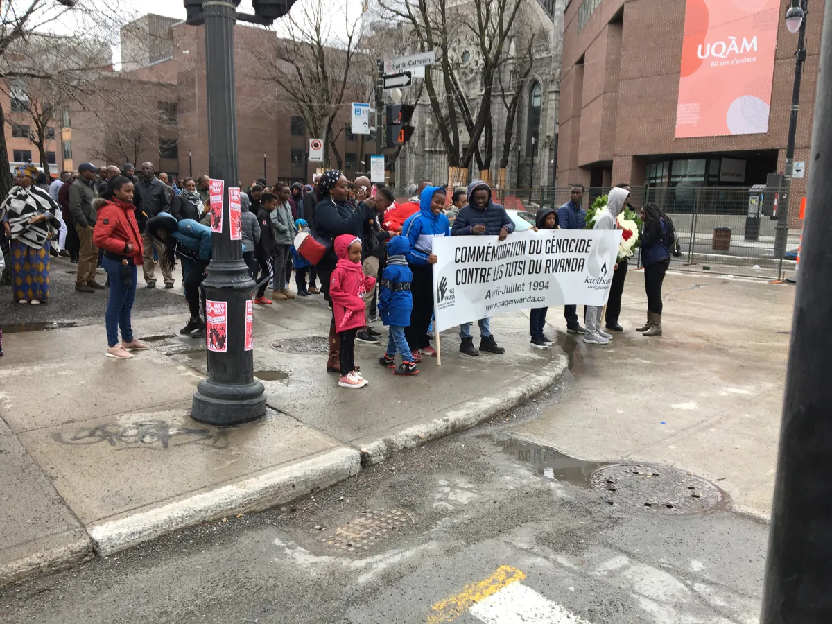 A group of people gathered on a street corner, holding a large banner reading, in French: “Commémoration du génocide contre les Tutsi du Rwanda, Avril-Juillet 1994, www.pagerwanda.ca”.