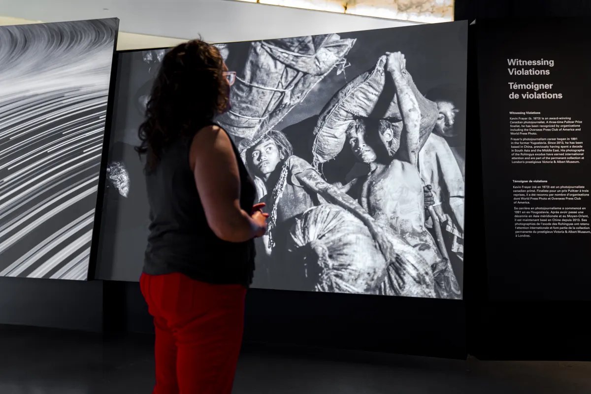 A woman looks at a large, projected photograph. Beside it, there is a text panel titled, "Witnessing Violations".