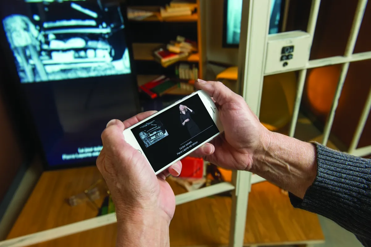 Two hands hold a smart phone in front of a museum exhibit. On the phone screen, a woman can be seen doing sign language interpretation. French subtitles are also visible.