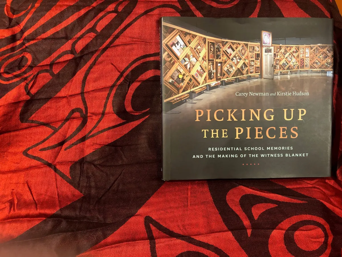 A book called “Picking up the Pieces: Residential School Memories and the Making of the Witness Blanket” lies on a red-and-black cloth. The cover includes a photograph of a large wood-framed art installation.