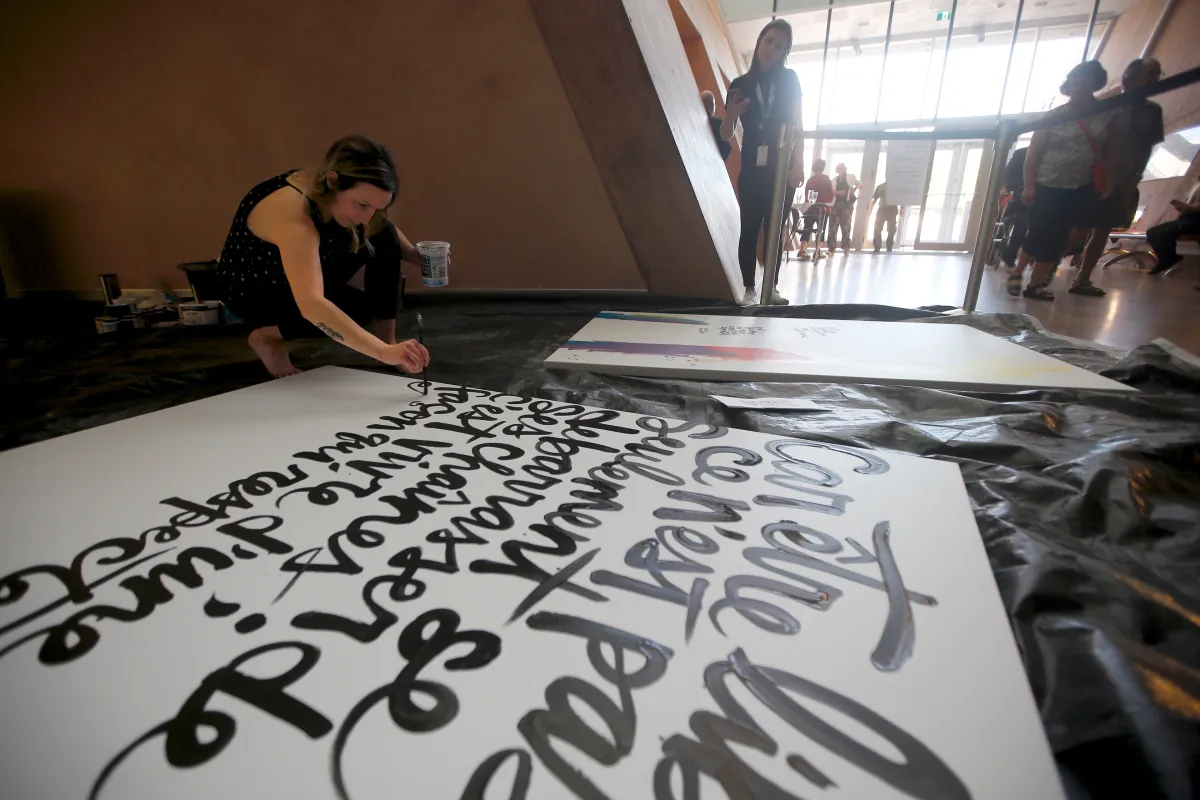 A woman crouches on the floor, painting artistic script on a large white canvas.