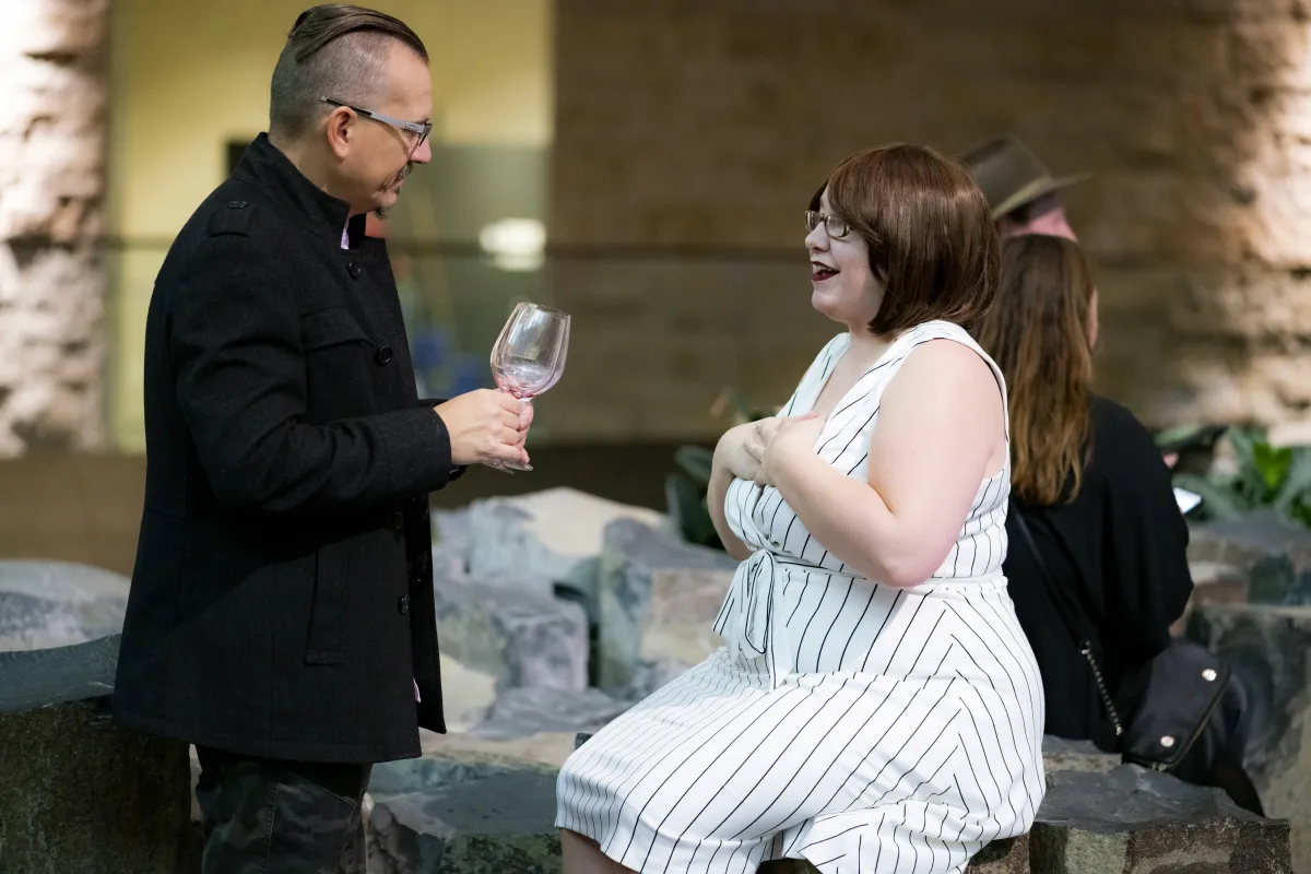 A couple in a Museum space. The woman sits on rock columns. The man is standing with a glass of wine.