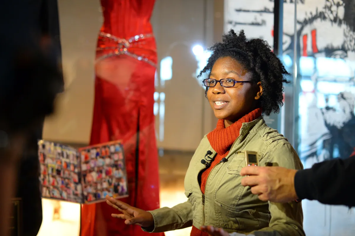 A smiling young woman stands in front of a red dress. She has a microphone on the lapel of her jacket. She is speaking to someone who is not seen in the photo.