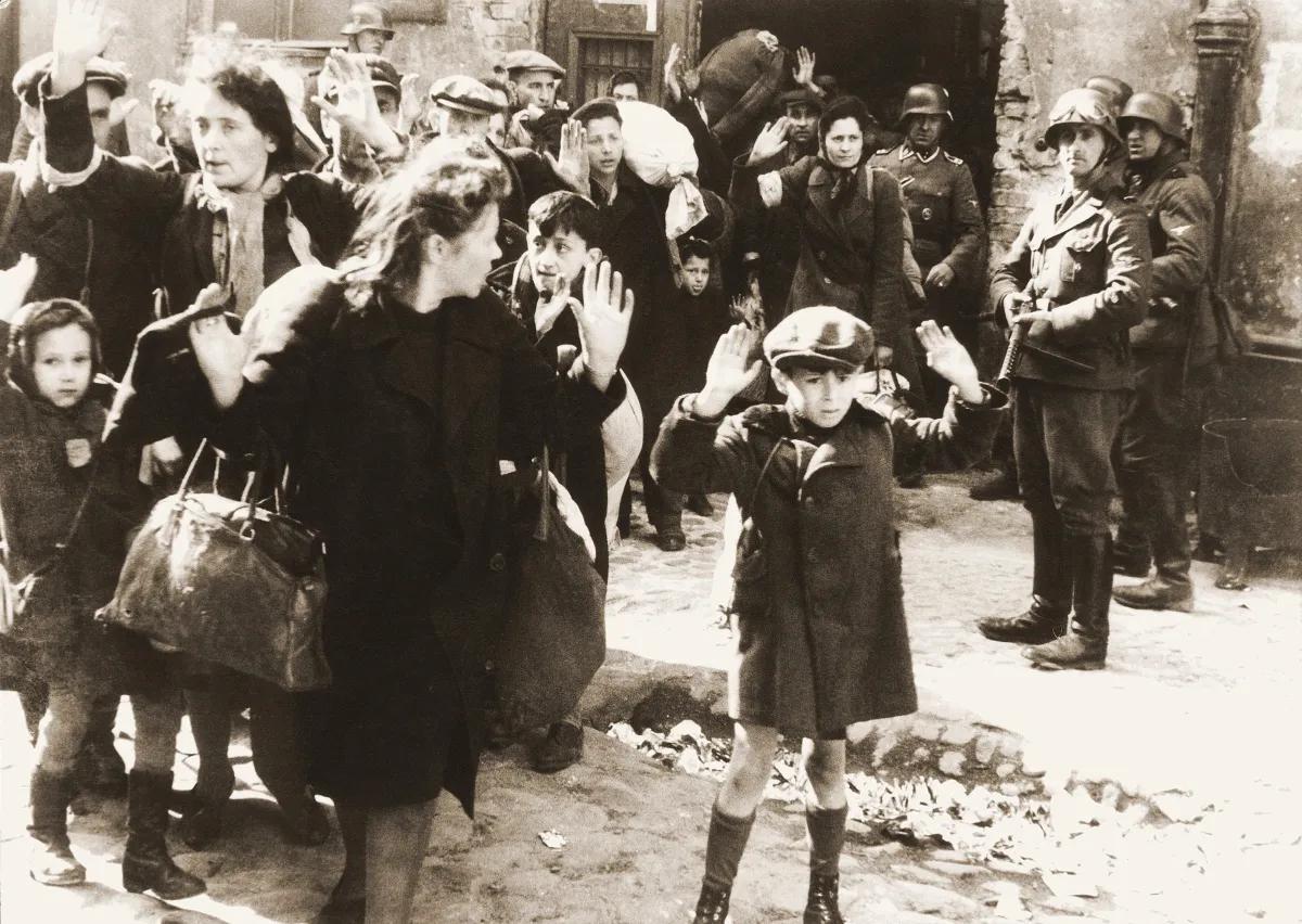 A black and white photo showing a group of people with their hands up being threatened by armed soldiers. A frightened-looking young boy is prominent in the foreground.