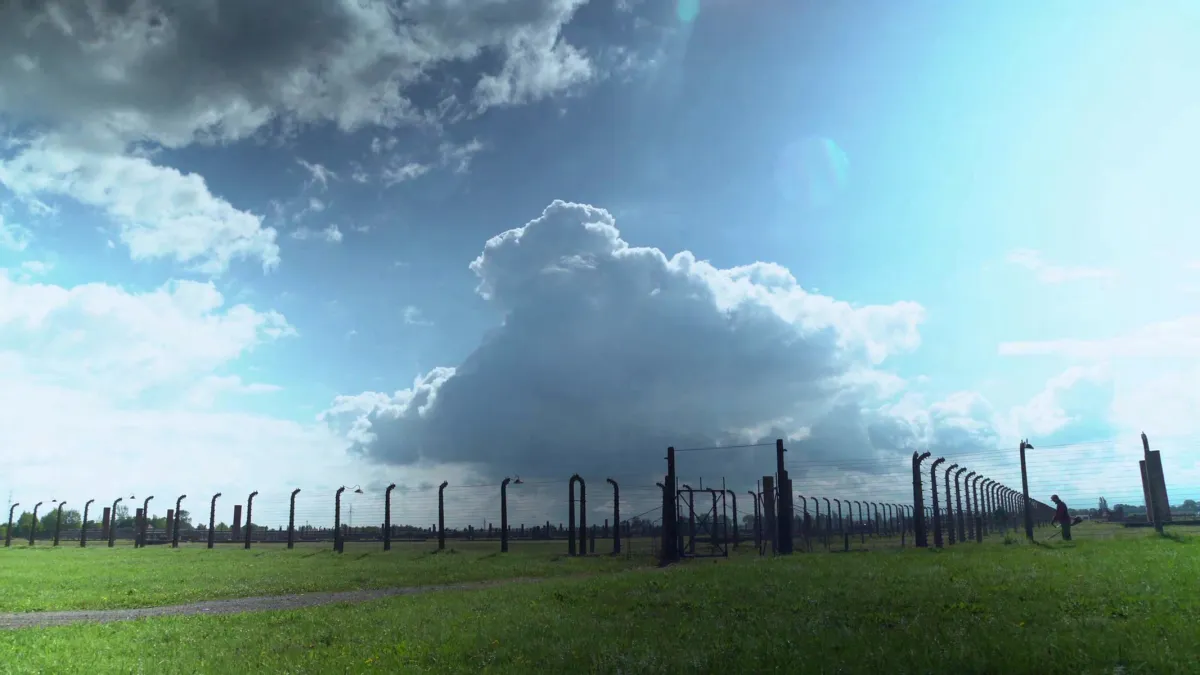 A tall, large black metal fence erected in a field of green grass. The sun shines as fluffy grey and white clouds float in the blue sky.