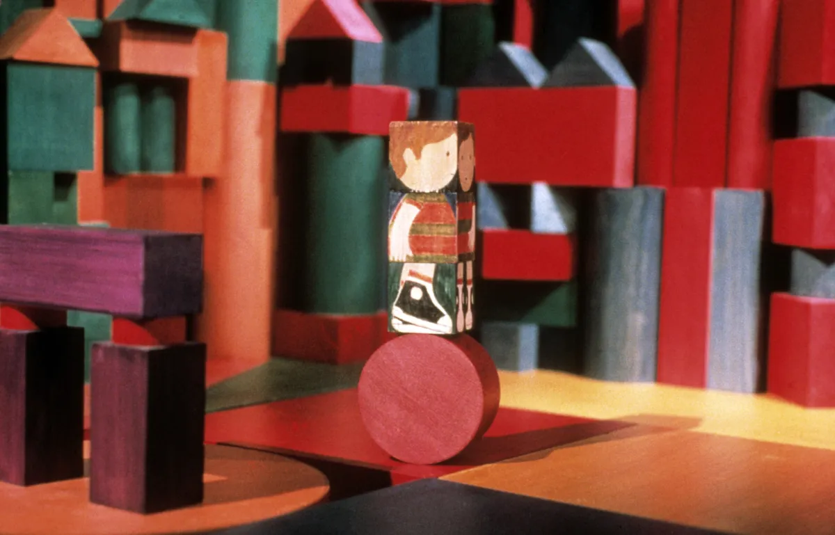 A wooden block painted with the image of a child, stands on top of a round block. Blocks of varying shapes, colours and sizes appear in the background.