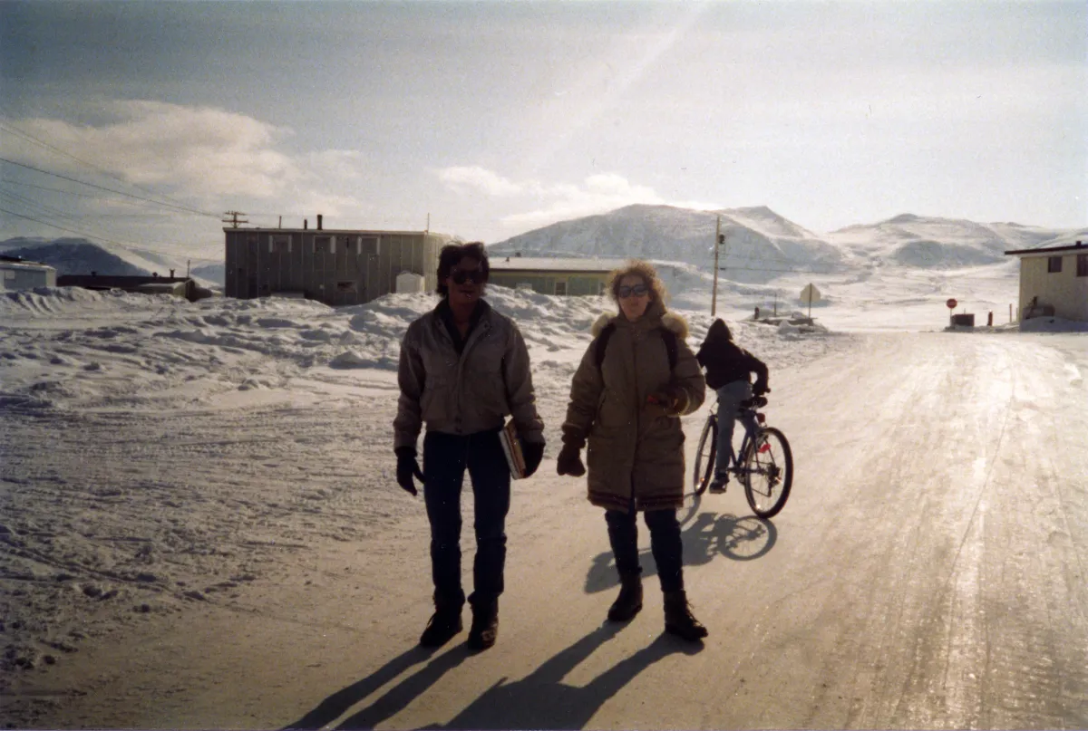 Two people in winter coats stand in a snowy landscape with hills and buildings in the distance.