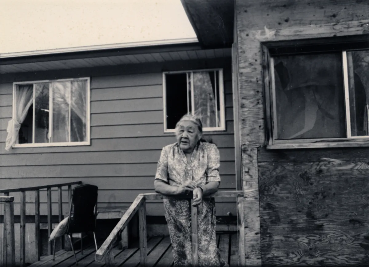 A white-haired woman leans on a porch railing outside a house.