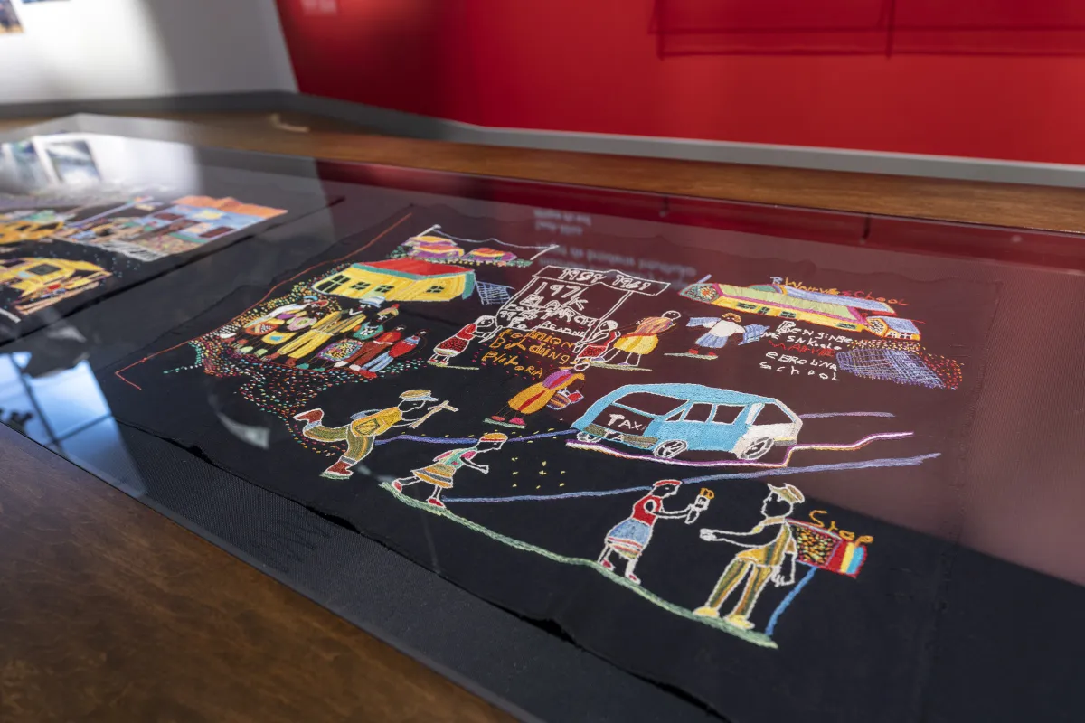 A colourful piece of embroidery on a black background depicts a scene with people, houses and vehicles.