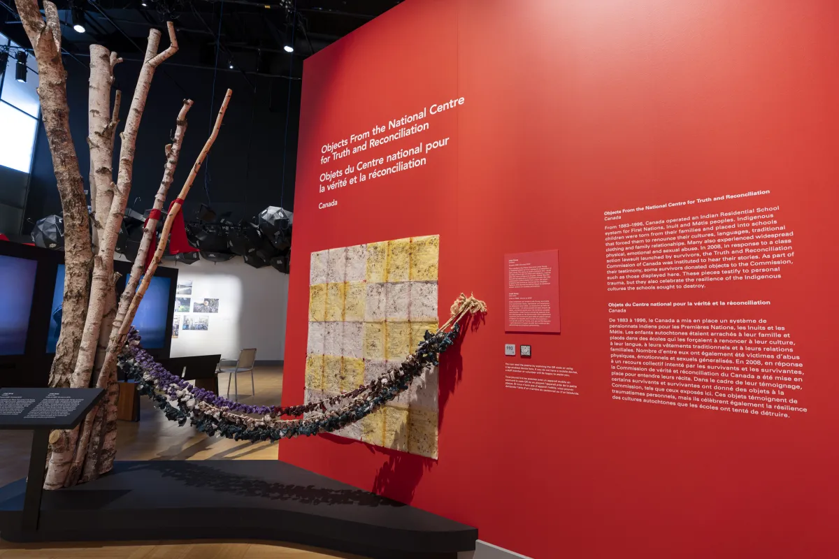 Ropes affixed with dozens of colourful cloth pouches are suspended between a birch tree and a red wall.