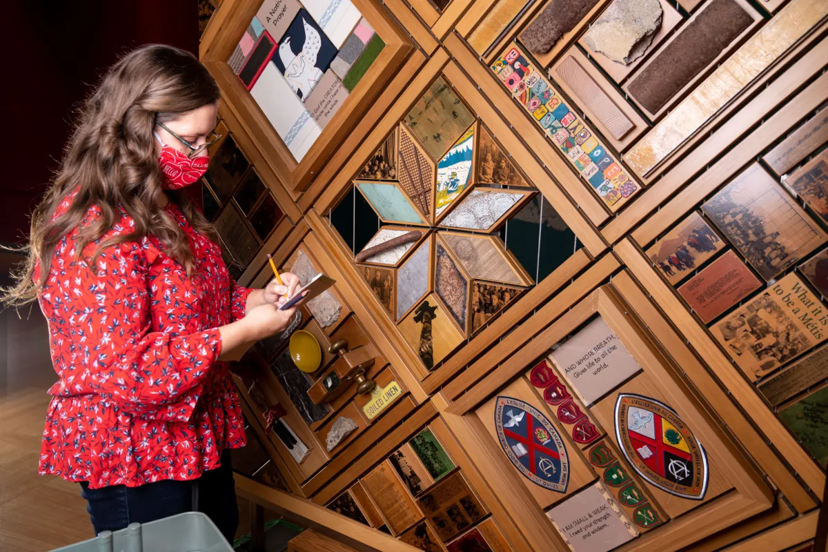A woman wearing a facemask writes in a notepad while standing in front of a large, wood-framed art installation that is full of objects.