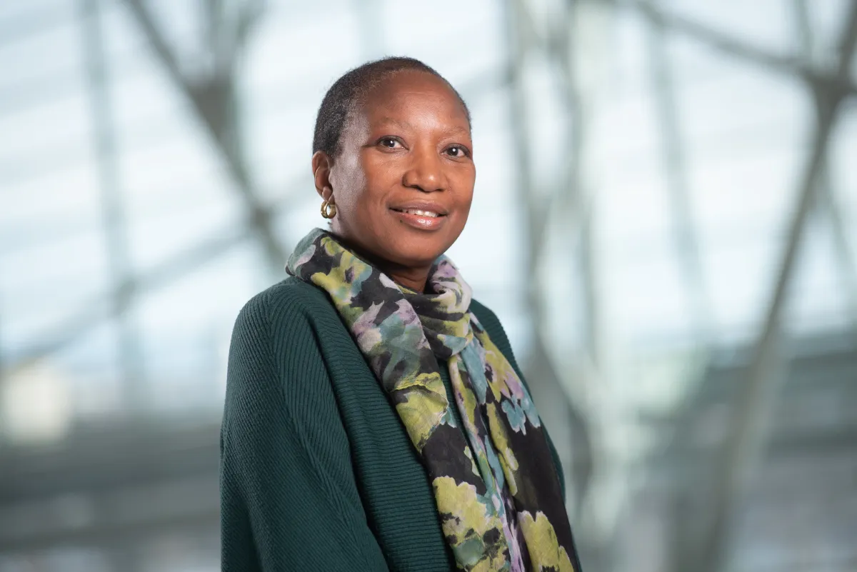 A black woman with short hair wears a dark green sweater with a green-purple-teal scarf. She's is standing at an angle and smiling at the camera.
