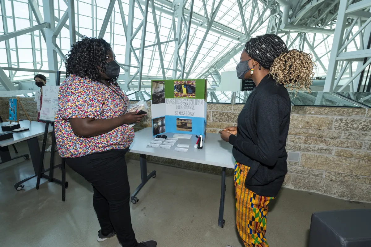 Two women speaking in between a poster that reads "Basics for the blind, Freetown, Sierra Leone"